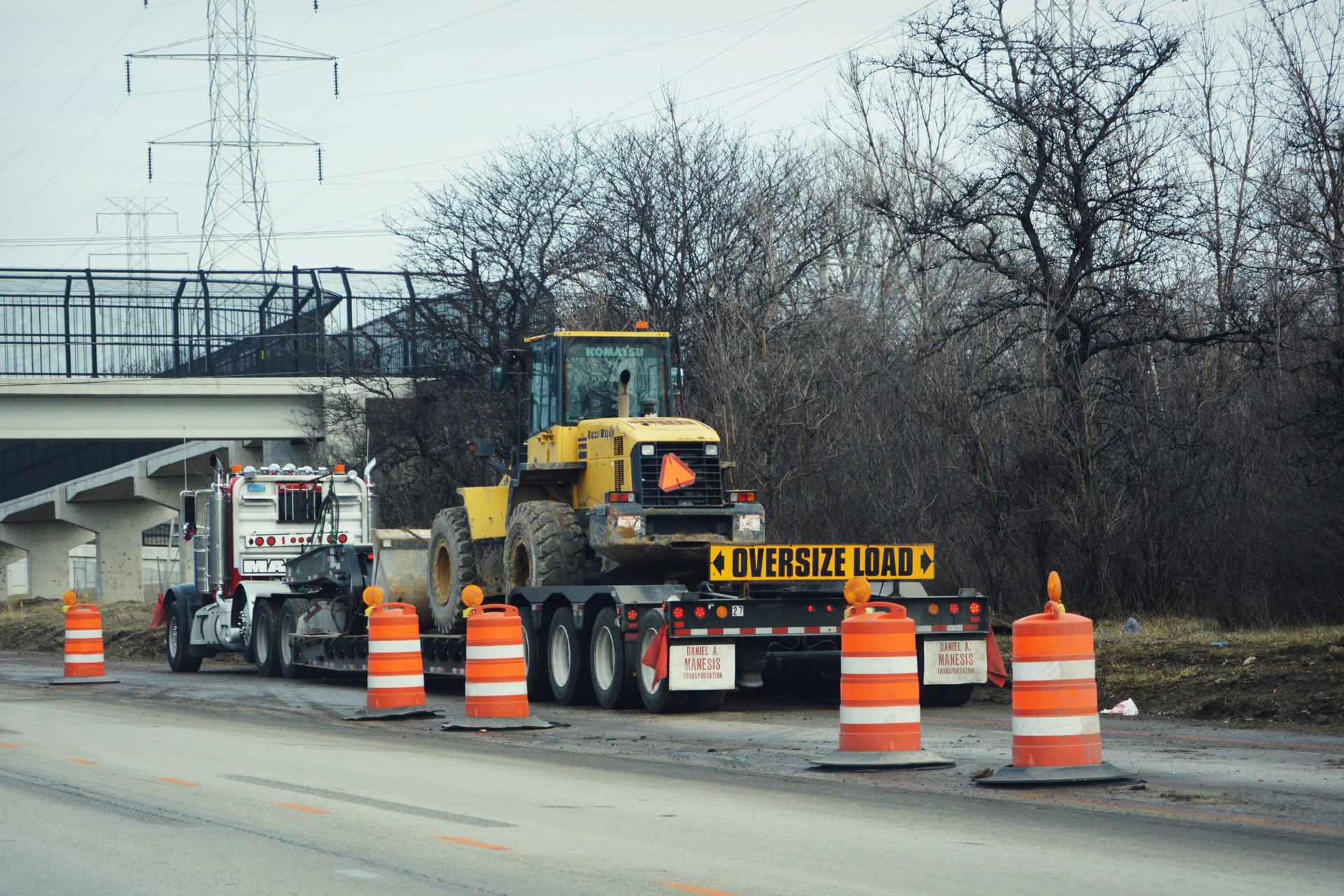A semi truck tractor bed pulling a yellow tractor with orange construction cones by a bridge on an interstate highway in Wisconsin.