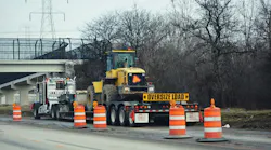 A semi truck tractor bed pulling a yellow tractor with orange construction cones by a bridge on an interstate highway in Wisconsin.