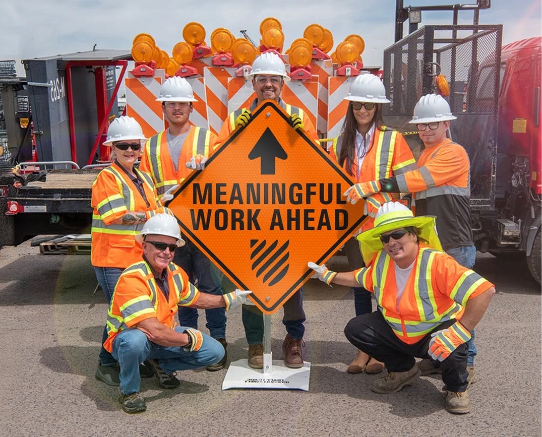 Road workers gathered around an orange construction sign stating 'Meaningful Work Ahead.'