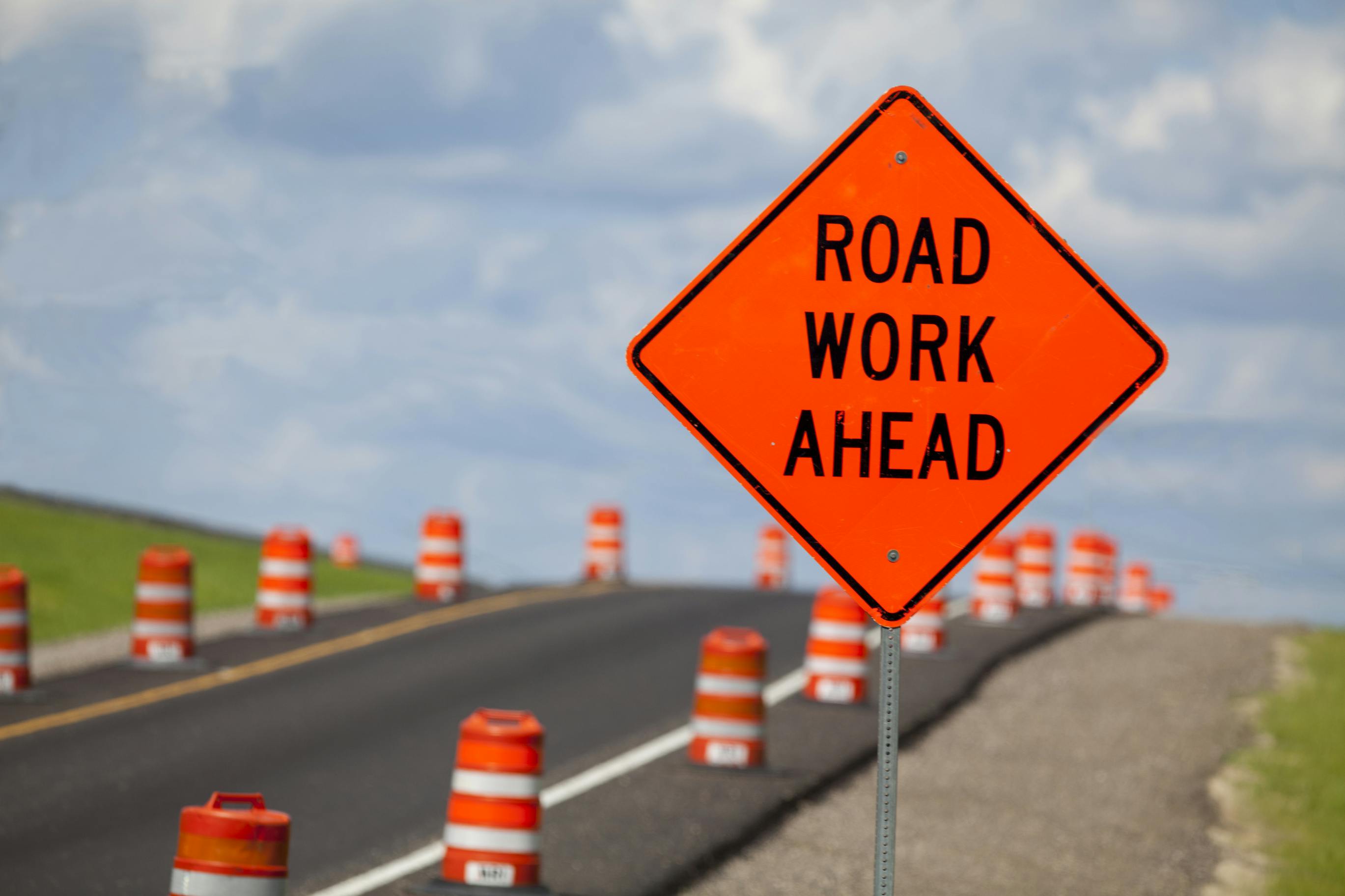 A road work ahead sign on the side of a roadway flanked by orange cones.