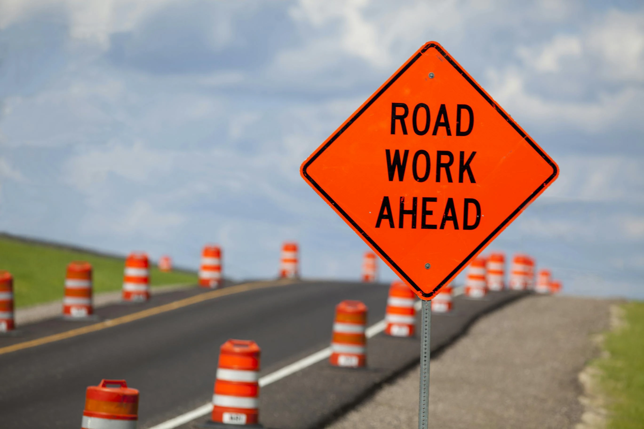 A road work ahead sign on the side of a roadway flanked by orange cones.